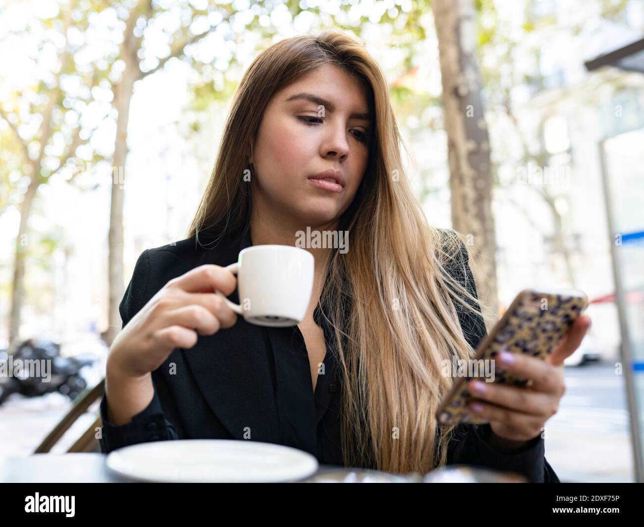 Woman using mobile phone while drinking coffee at sidewalk cafe in city Stock Photo