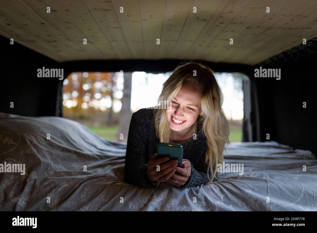 Smiling woman with blond hair lying on bed while using smart phone in camper van Stock Photo