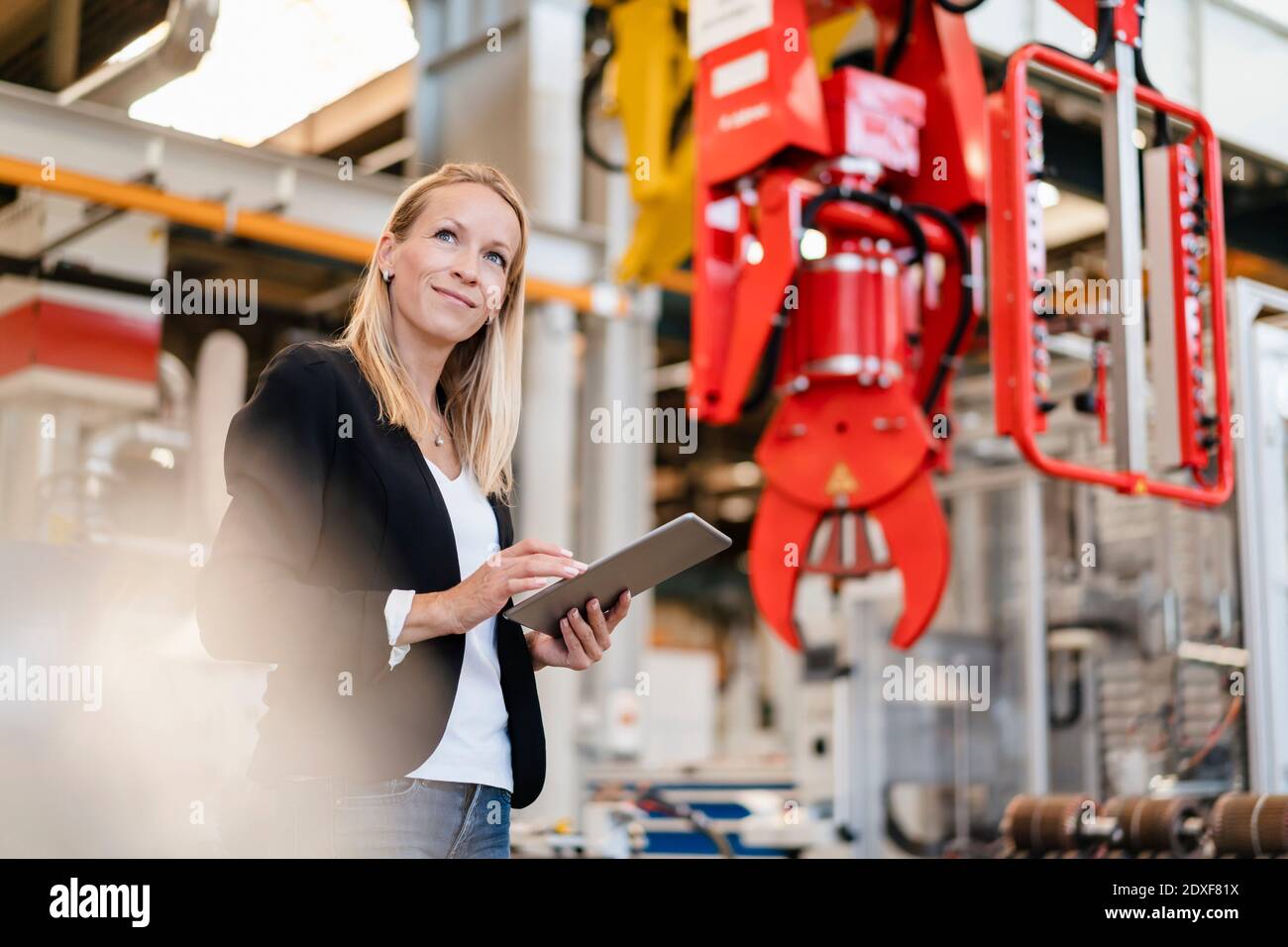 Smiling blond businesswoman looking away while holding digital tablet standing in factory Stock Photo