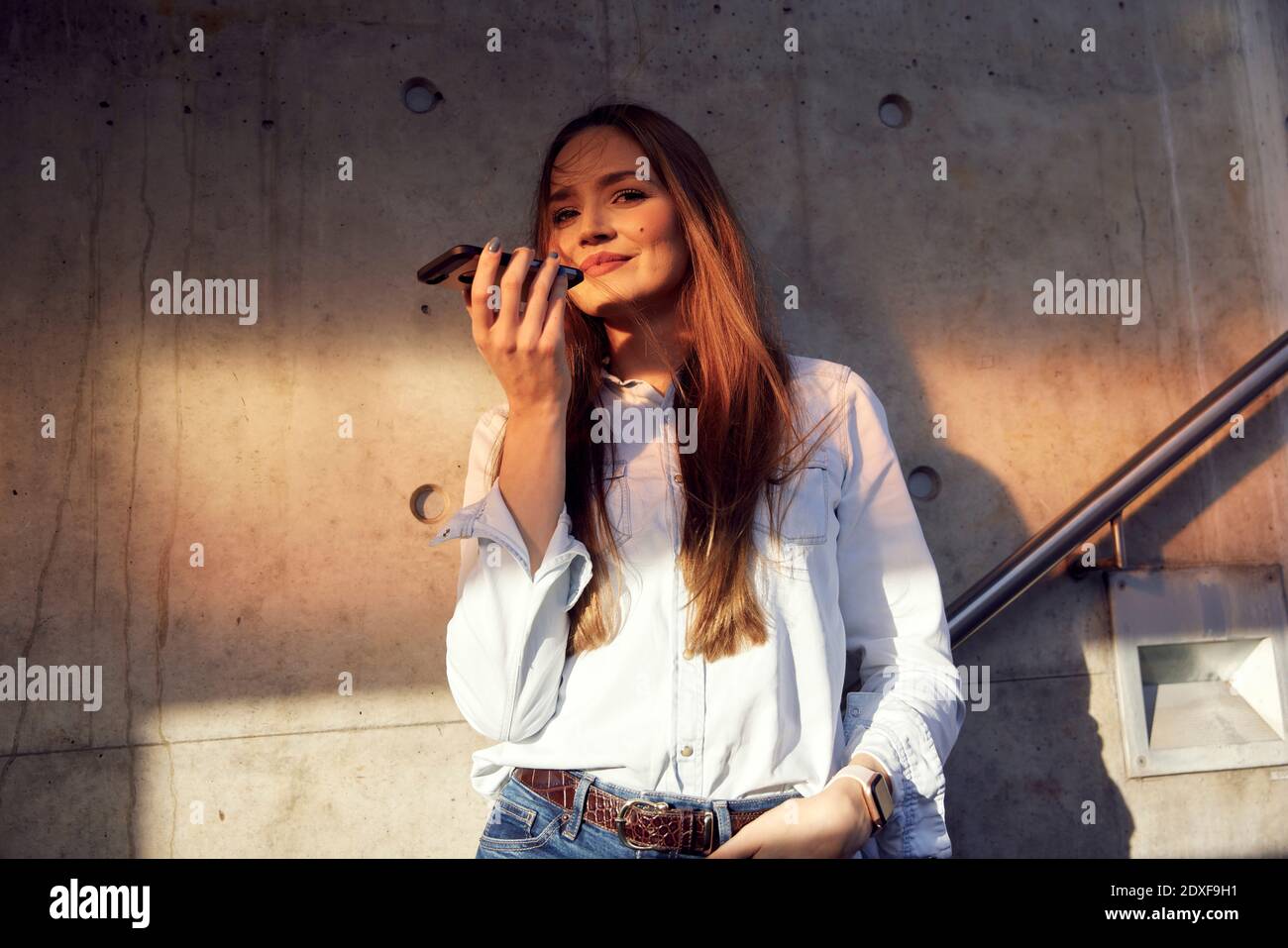 Young businesswoman with hands in pockets talking on mobile phone while standing against wall Stock Photo