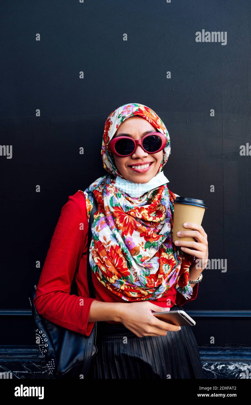 Smiling Muslim woman holding mobile phone and coffee cup against black wall during COVID-19 Stock Photo
