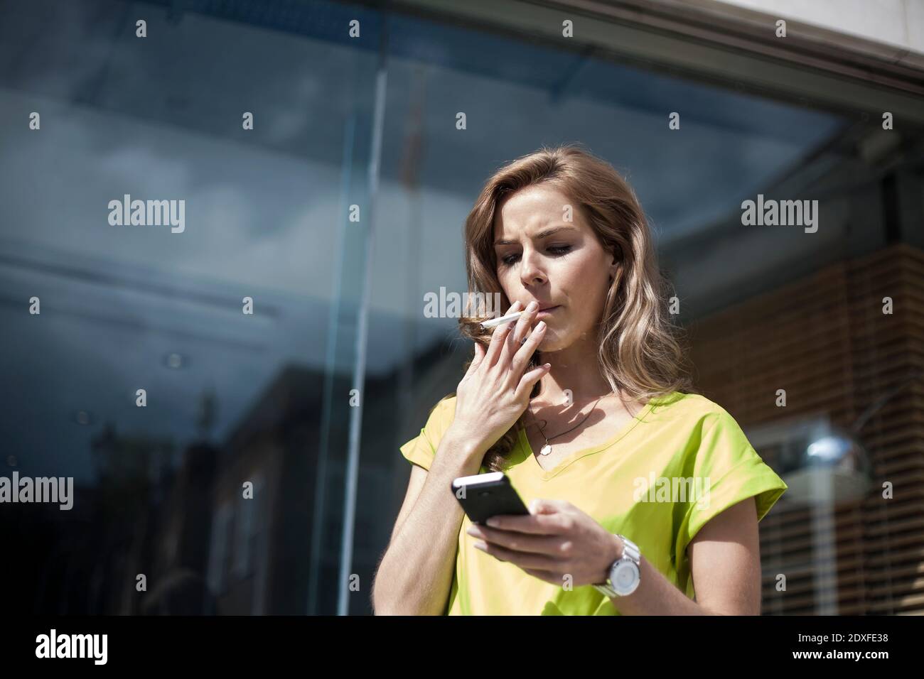 Young female entrepreneur using smart phone while smoking against office building on sunny day Stock Photo