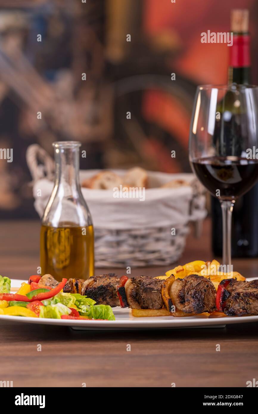 roasted beef skewer with cut vegetables and french fries on a plate, with out of focus bottle of oil and glass of wine at the back. Selective focus. Stock Photo