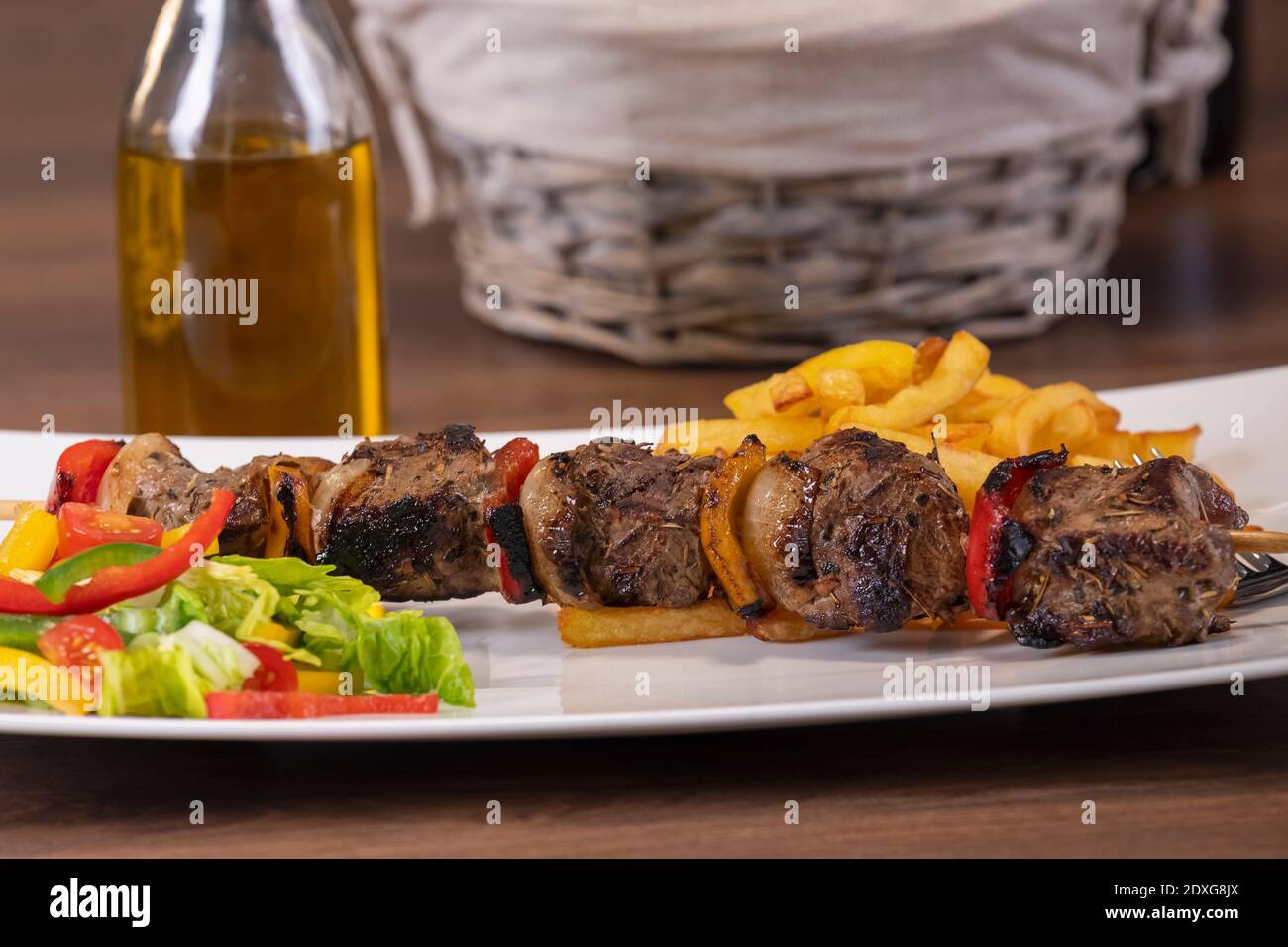 Juicy roasted beef skewer with freshly cut vegetables and french fries on a plate with an out of focus bottle of oil at the back. Selective focus. Stock Photo