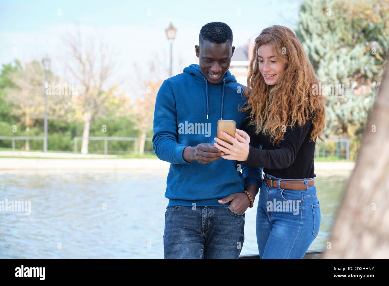 African black man and redhead caucasian woman smiling and looking at their smartphone in a park. Young multiracial couple portrait. Stock Photo