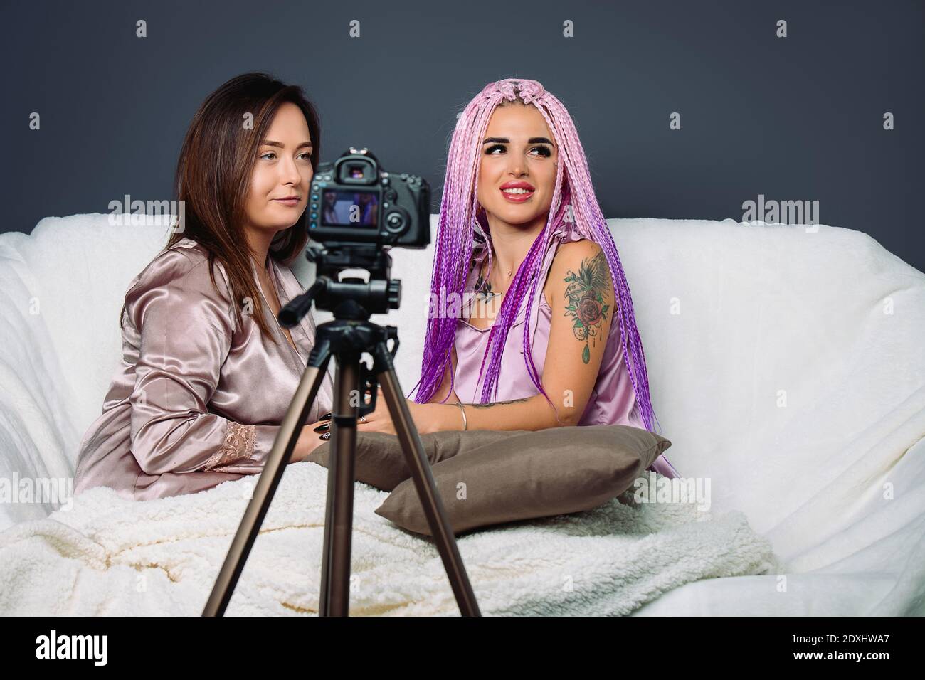 Fashion girlfriends are looking out, smiling on the sofa with pillows. Online photo camera in studio Stock Photo