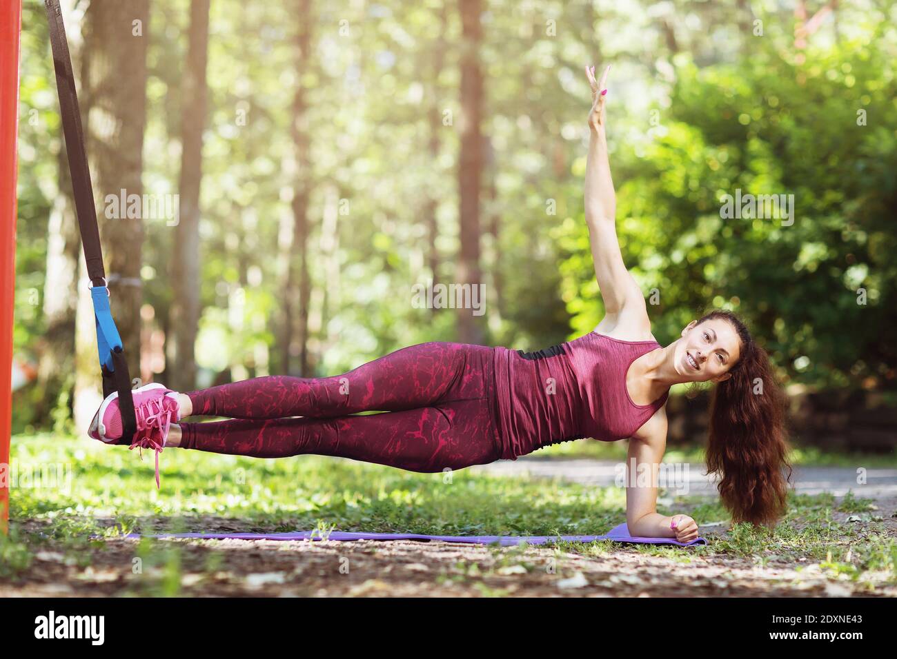 A young woman performs a side plank exercise on a suspended machine attached to a horizontal bar in a park. Stock Photo