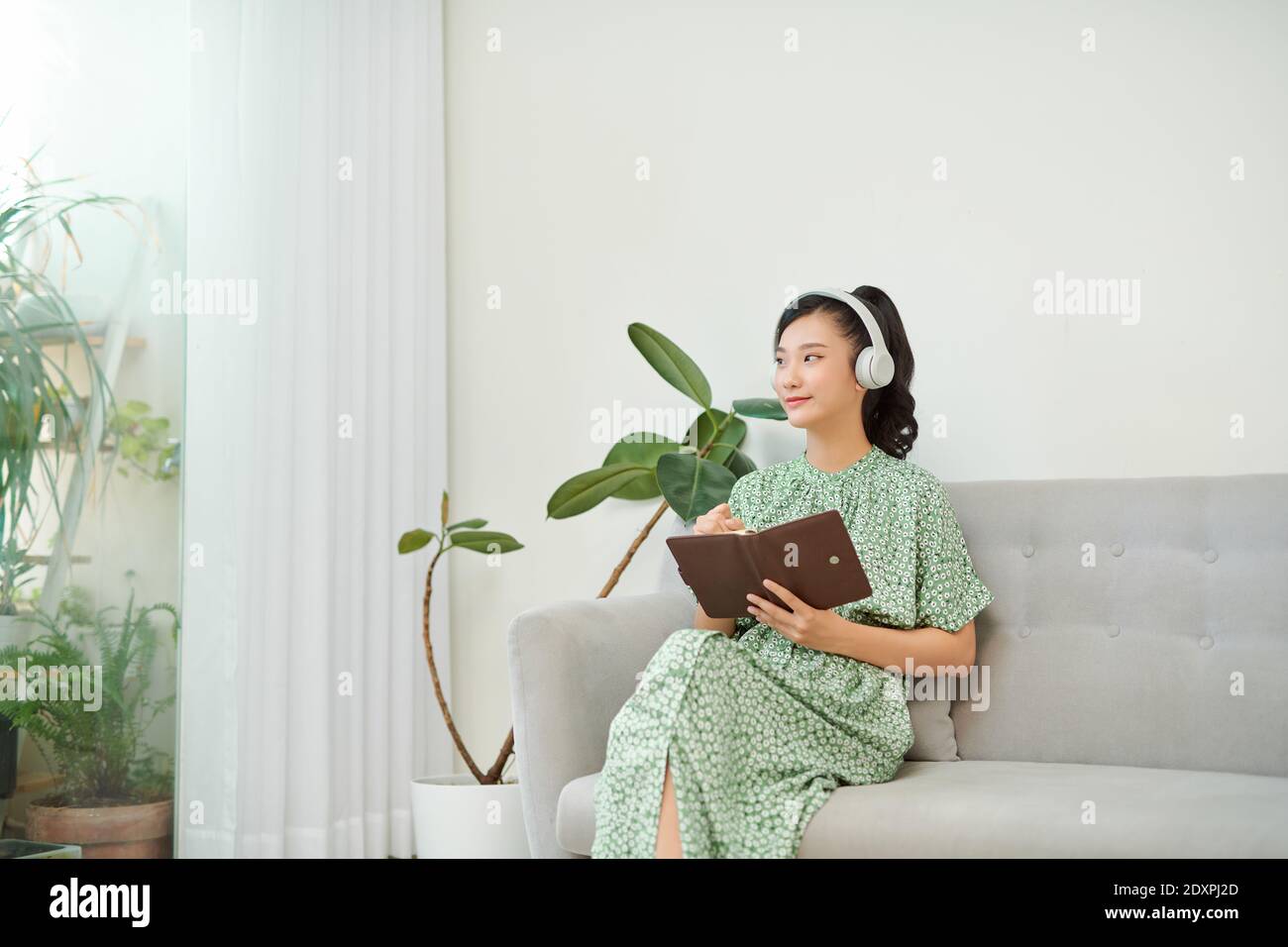 mage of focused asian woman using headphones and writing down notes while sitting on sofa at home Stock Photo