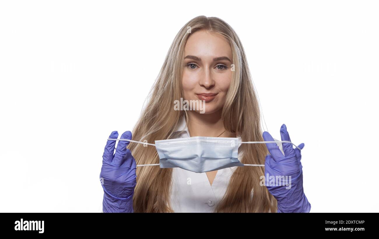 About to put on medical face mask young nurse with long straight hair looking at camera. Caucasian woman wearing white medical uniform isolated on Stock Photo
