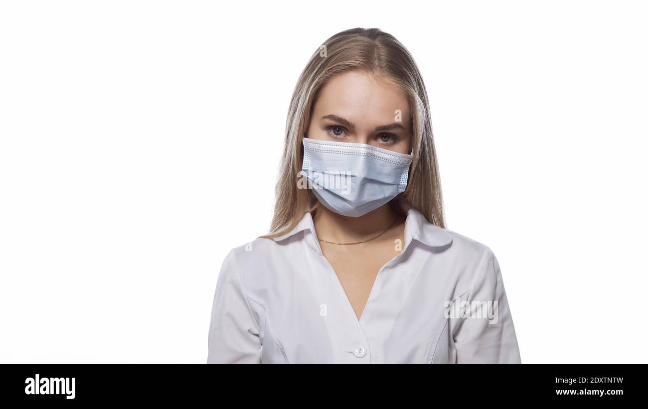 Looks sullenly young nurse in a medical mask and white uniform with blond straight hair looking at the camera. Isolated on white background Stock Photo