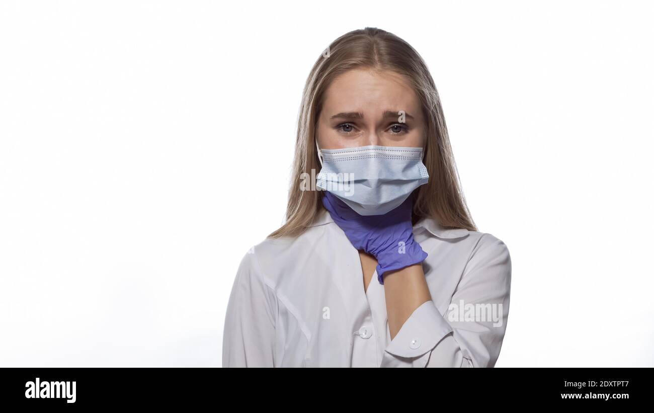 Sore throat nurse in a medical mask and disposable gloves touching her throat, looking at camera with pain in her eyes. Isolated on white background Stock Photo