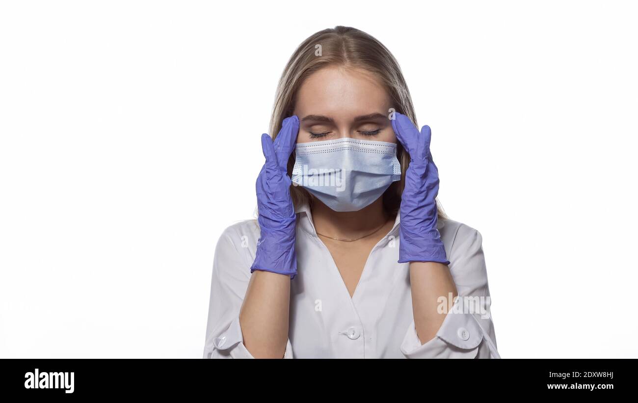 Suffering headache nurse in a medical mask and disposable gloves at work, massaging the temporal lobes of the head, with eyes shut. Isolated on white Stock Photo