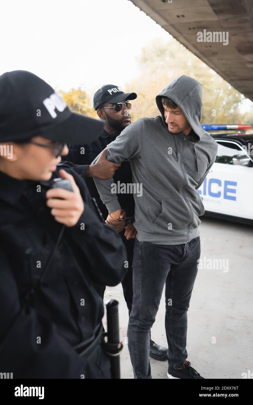 african american policeman handcuffing hooded offender with blurred colleague on foreground outdoors Stock Photo