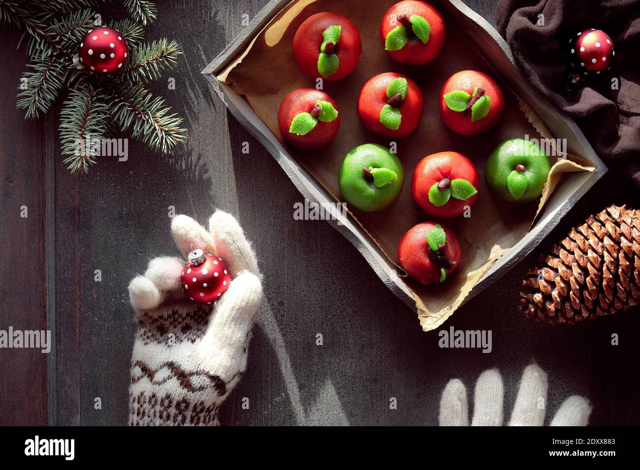 Marzipan sponge apples. Tasty creative dessert on rustic wooden tray with textile cotton towel. Hands in wool gloves. Stock Photo