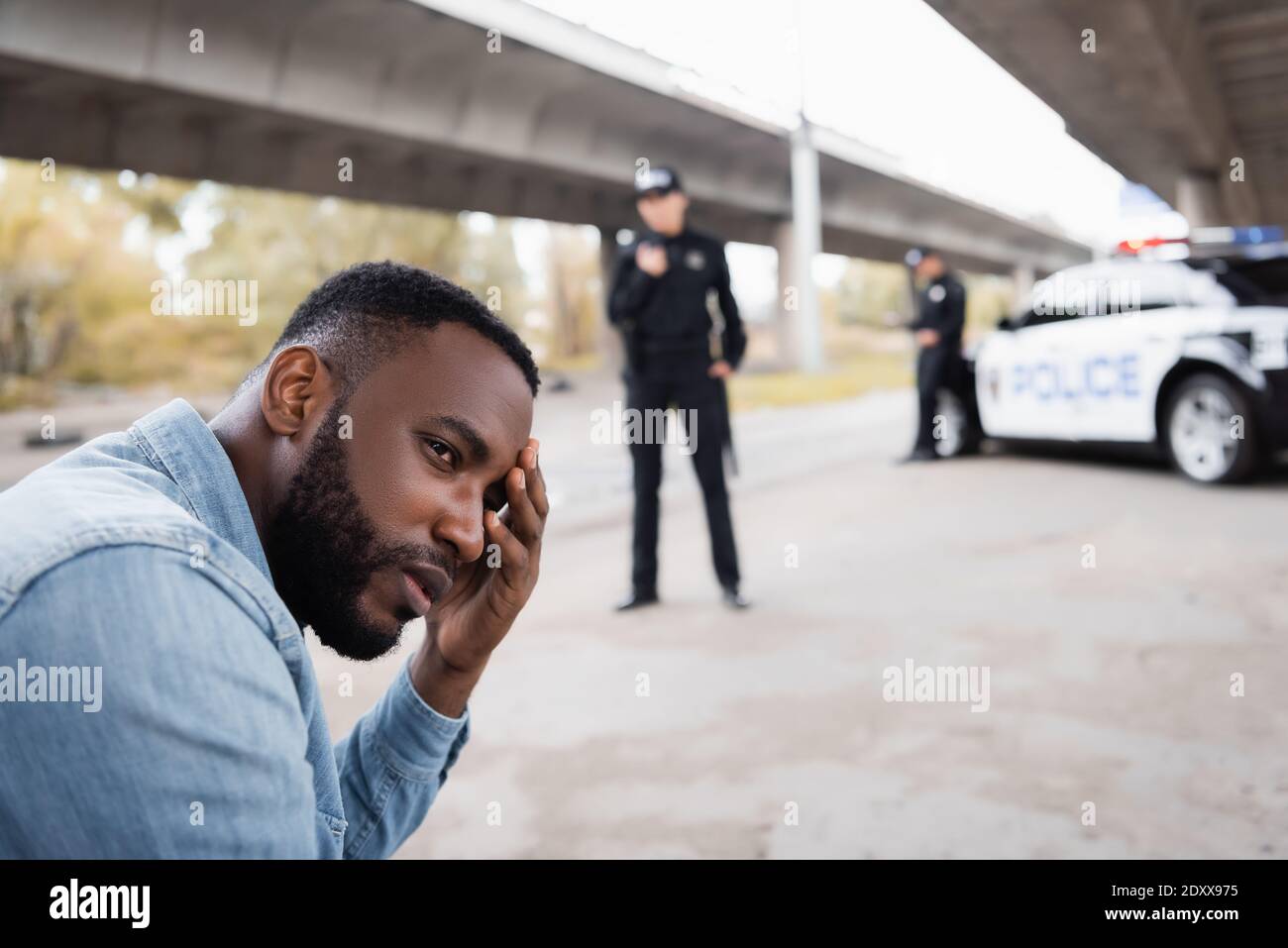 Upset african american victim looking away near police officers and car on blurred background on urban street Stock Photo