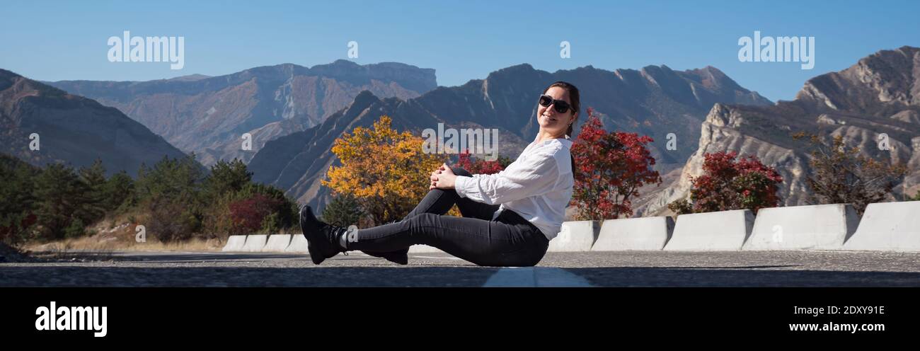 Young woman in white hoodie and sunglasses sits on grey asphalt road barrier against red and orange trees and rocky mountains on autumn day Stock Photo