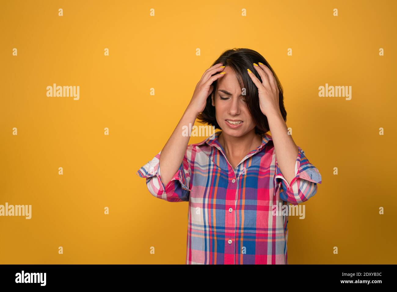 Having a headache pretty young girl holding a head with two hands and grimace of pain on the face wearing plaid shirt isolated on yellow background Stock Photo