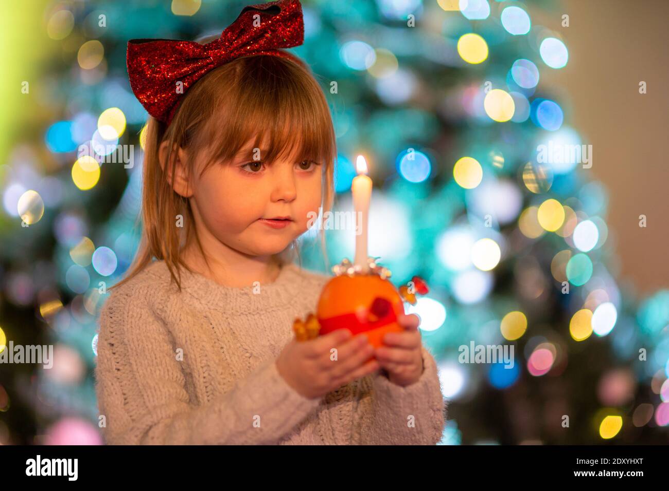 Cradley Heath, West Midlands, UK. 24th Dec, 2020. Four-year-old Hermione Hadlington stands proudly with her Christingle orange during the Christmas Eve service for children at Holy Trinity church, Cradley Heath, West Midlands. In the Christingle service the orange represents the world, the red ribbon symbolises the love and blood of Christ, the sweets represent all of God's creations, and the lit candle represents Jesus's light in the world, bringing hope to people living in darkness. Credit: Peter Lopeman/Alamy Live News Stock Photo