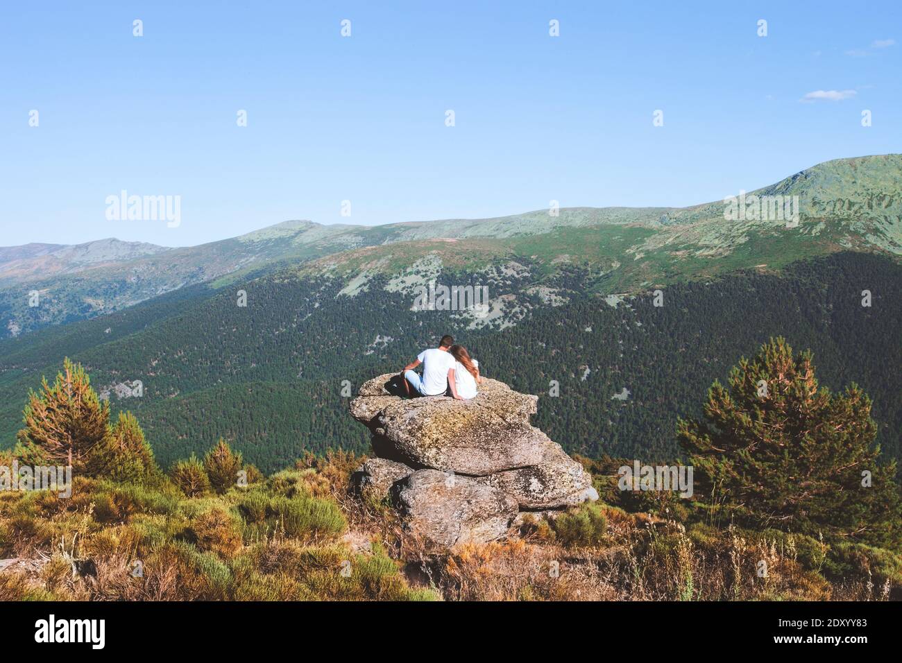 Couple in love is sitting on a high rock looking at the mountain valley on a sunny summer day Stock Photo