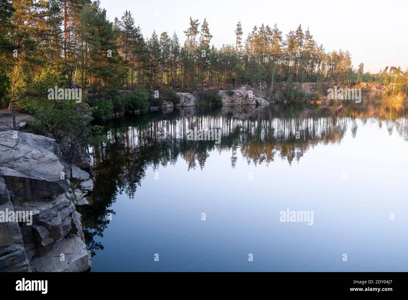 View of a beautiful clear lake among granite rocks and pine forest reflected in the water at sunset in summer Stock Photo