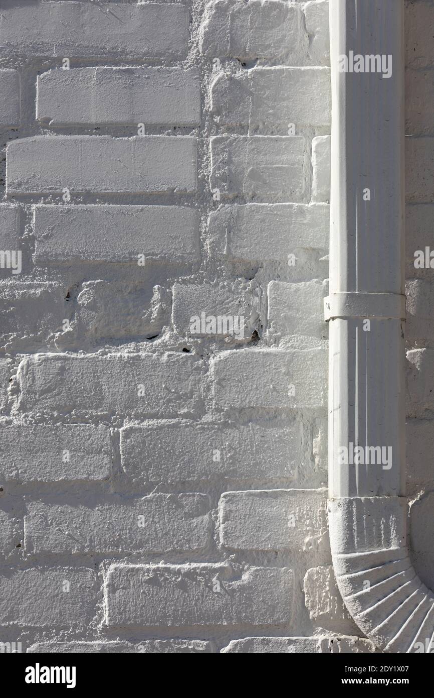 Vertical close-up of a white brick wall, a metal downspout at right, both painted white, all in bright side light. Stock Photo