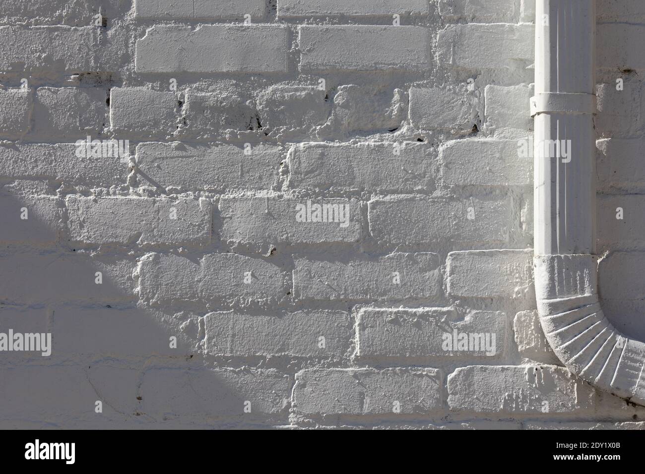 Horizontal close-up of a white brick wall, a metal downspout at right, both painted white, all in bright side light. Stock Photo