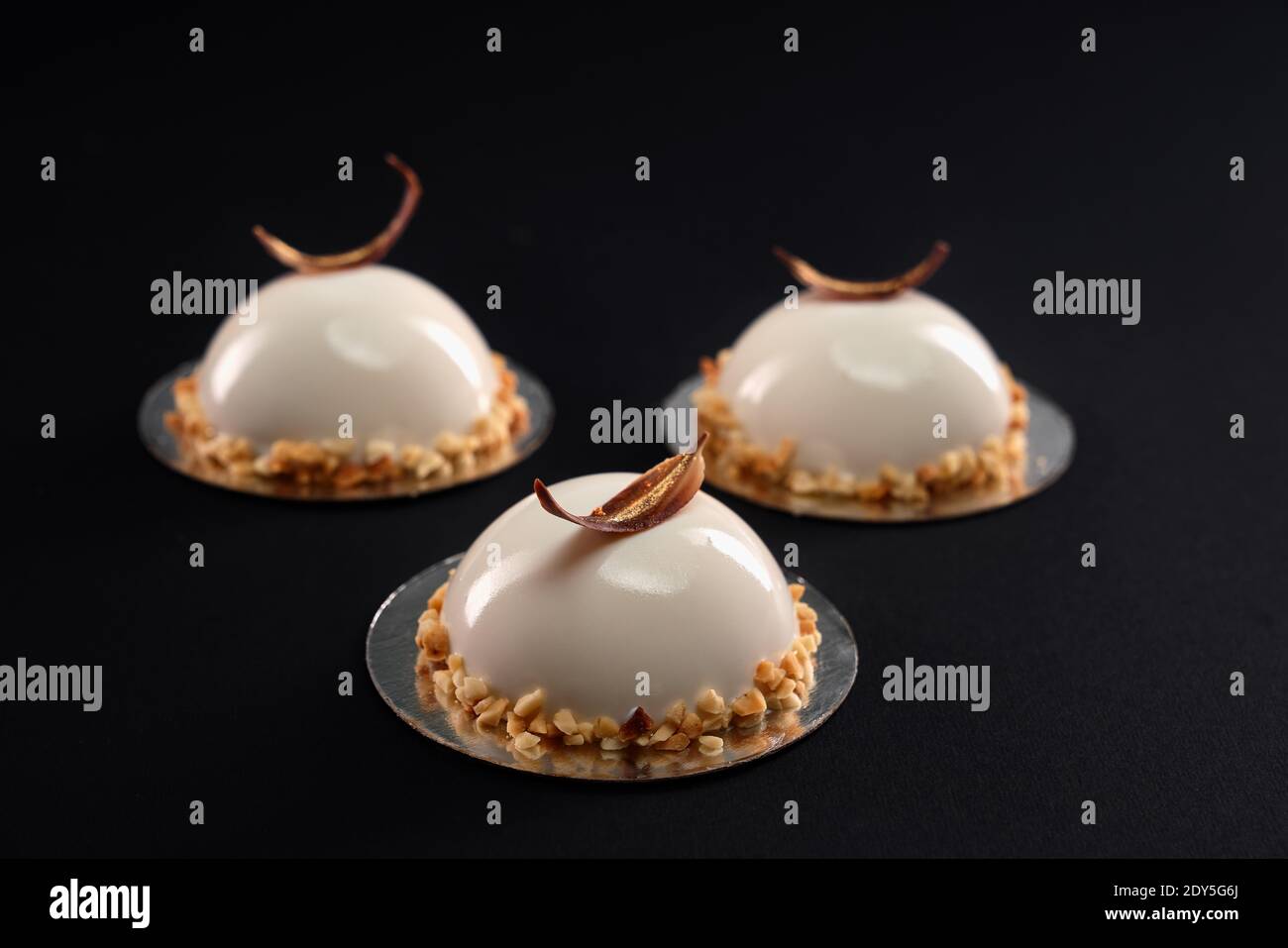 White half sphere cakes decorated with nuts and chocolate feather isolated on black background. Dessert with smooth surface, mousse and mirror glaze. Stock Photo