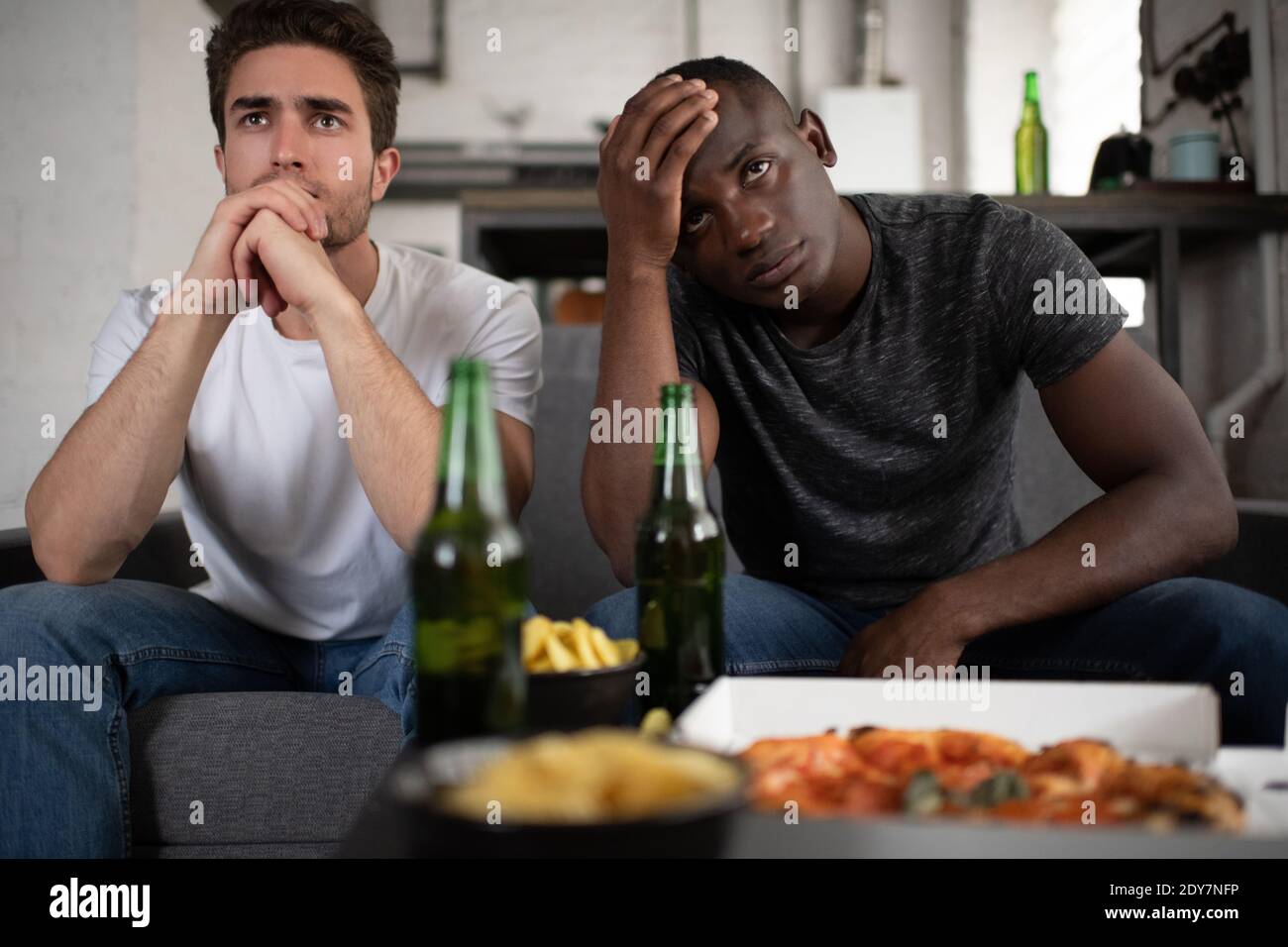 Displeased multicultural men in casual outfit clasping hands and touching forehead while sitting on sofa near table with snacks and beer and watching Stock Photo