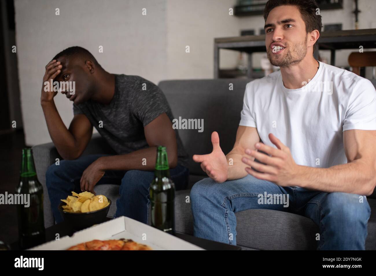Angry man gesturing and swearing while sitting on couch near table with beer and snacks while watching lame football match with frustrated black frien Stock Photo