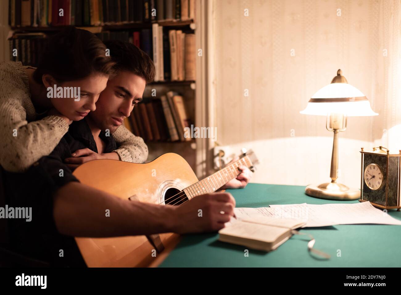 Young woman hugging focused boyfriend and helping to create song in evening in dark vintage apartment Stock Photo