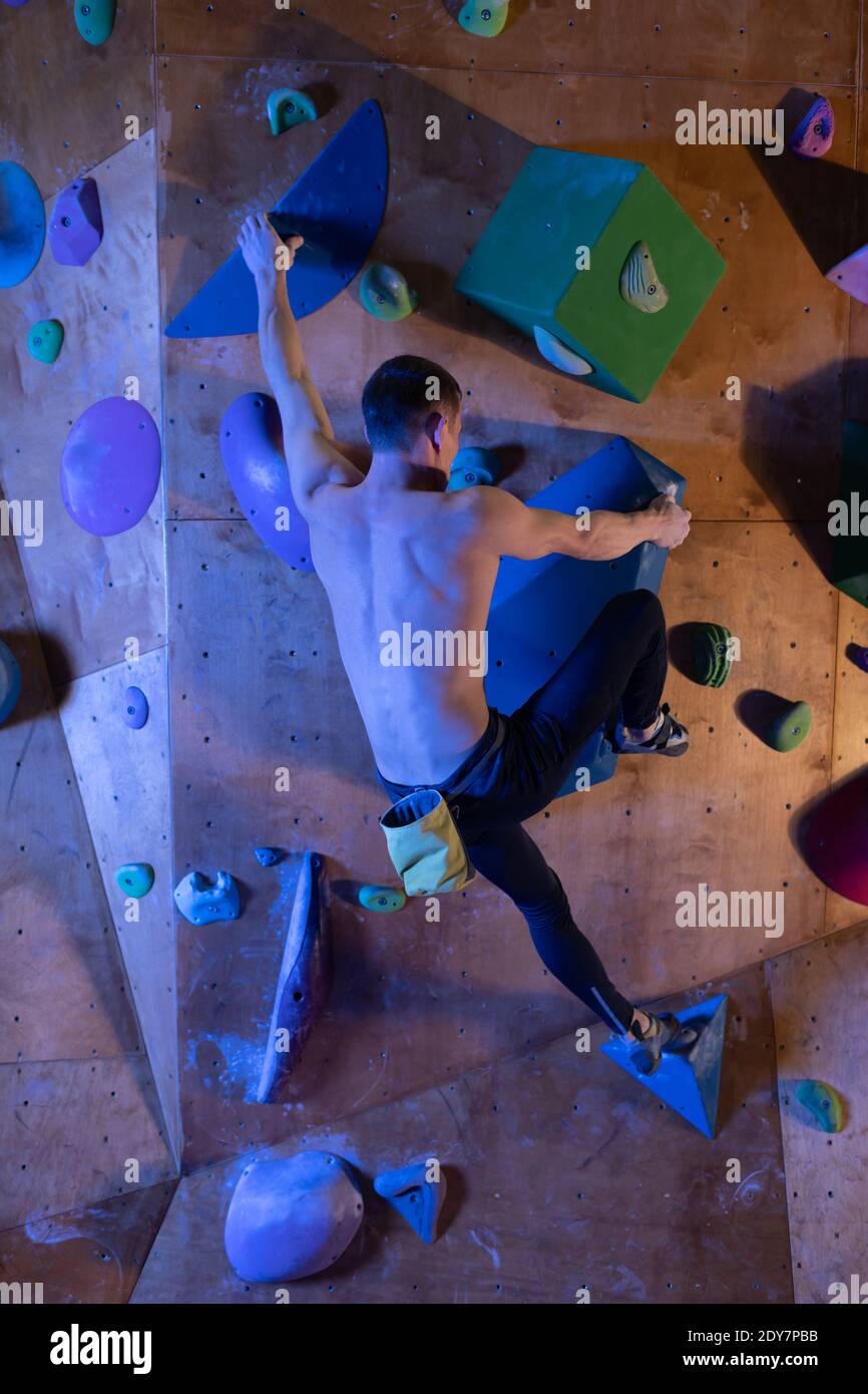 Back view of shirtless muscular man climbing wall without belay during bouldering workout in
