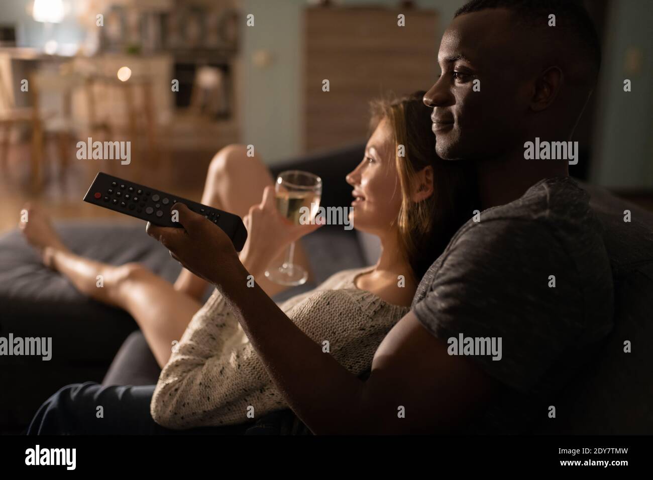 Black man changing channels on TV and embracing girlfriend with wine while relaxing on sofa at home Stock Photo