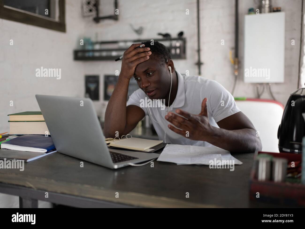 African American man facepalming in frustration while sitting at kitchen table and discussing assignment with classmates during remote lesson at home Stock Photo