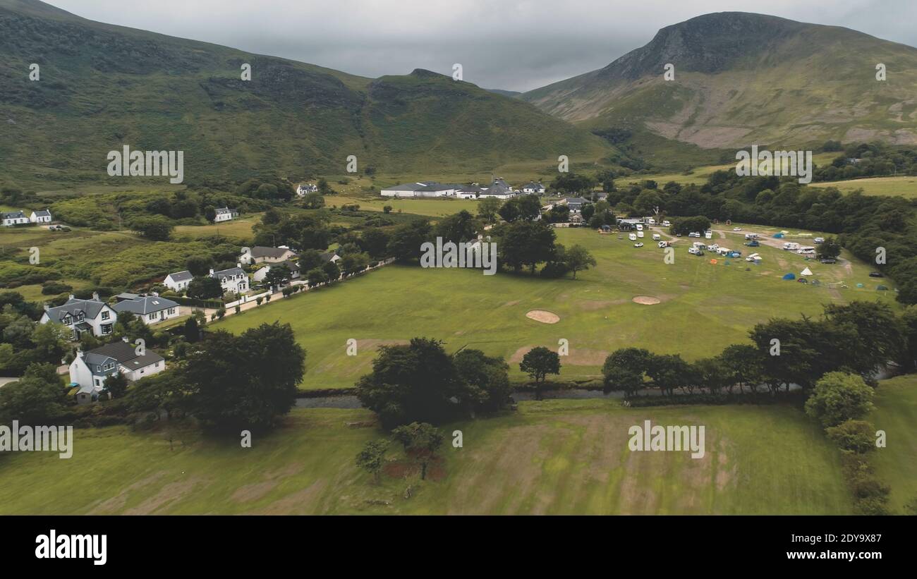 Whiskey distillery at mountain green valley. Nobody nature landscape at summer cloudy day. Scottish village cottages, houses at road aerial. High mounts of Arran Island, Scotland. Cinematic shot Stock Photo