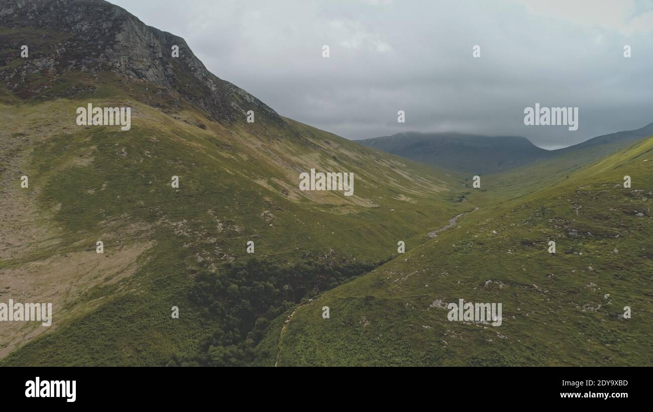 Green grass mountain ranges aerial. Path at greenery grassy mounts. Nobody nature landscape of Arran island, Scotland Grey clouds over top at summer cloudy day. Wild untouched scape at drone shot Stock Photo