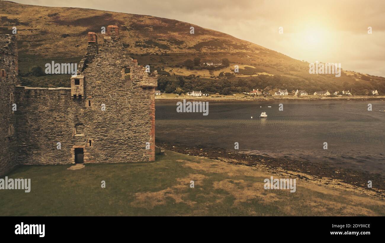 Closeup castle walls ruins at sun seascape aerial. Historic ruined palace on ocean coast. Green grass valley and mountain on sea shore. Ship on harbor of Loch-Ranza Bay, Arran island, Scotland, Europe Stock Photo