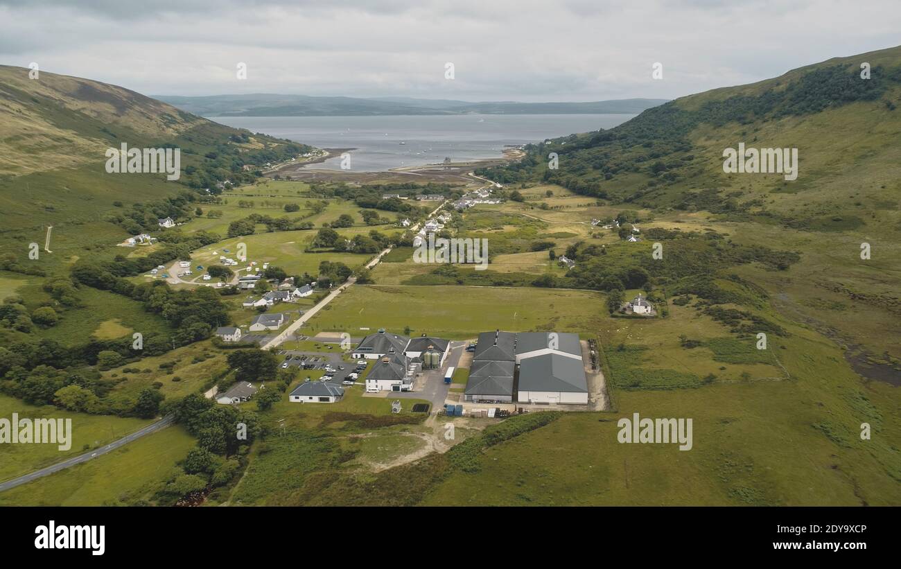 Scottish whisky distillery at village aerial. Road, cottages, houses at green mountain valley. Summer cloudy day. Nobody nature landscape of Arran Island, Scotland. Cinematic drone shot Stock Photo