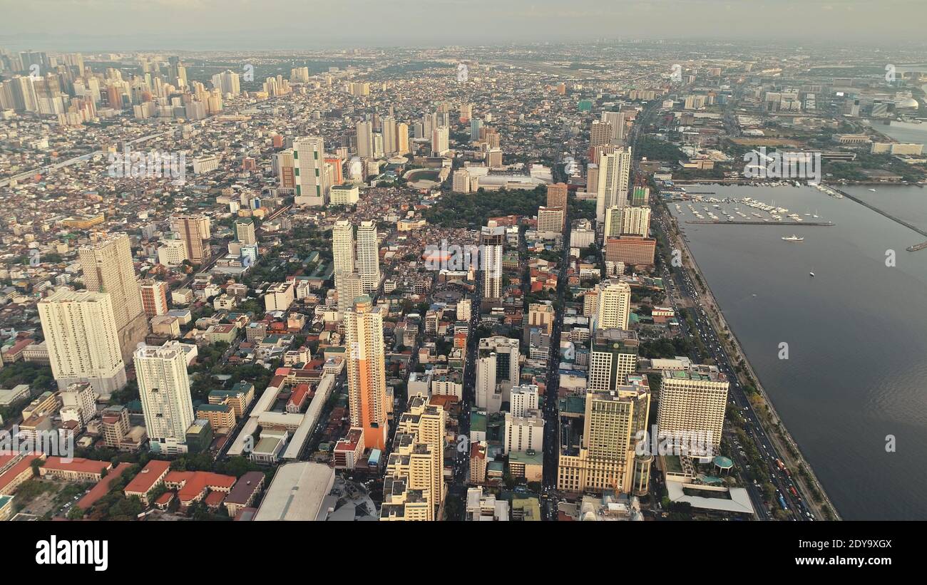 Metropolis port city of Manila at sea bay aerial view. Urban cityscape of streets: roads with multi-storey buildings, cottages, houses. Misty haze over town at summer dusk day. Cinematic panorama shot Stock Photo