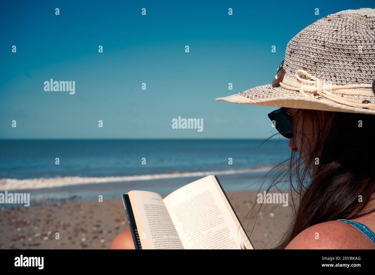 A dark-haired female in a Panama hat reading on the beach, beautiful sea view in the background Stock Photo
