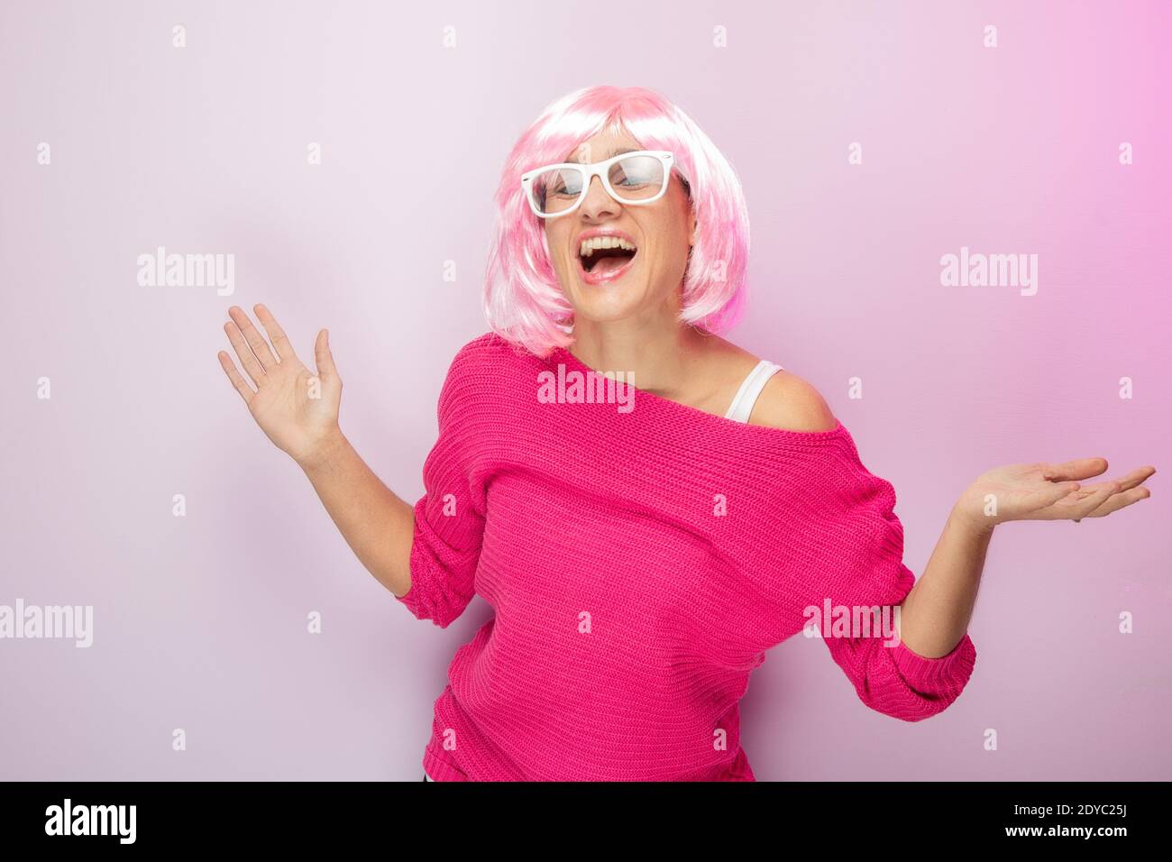 Caucasian woman with pink wig and sweater, wearing white glasses, posing funny at the photo studio. Funny expressive women and colorful portrait conce Stock Photo