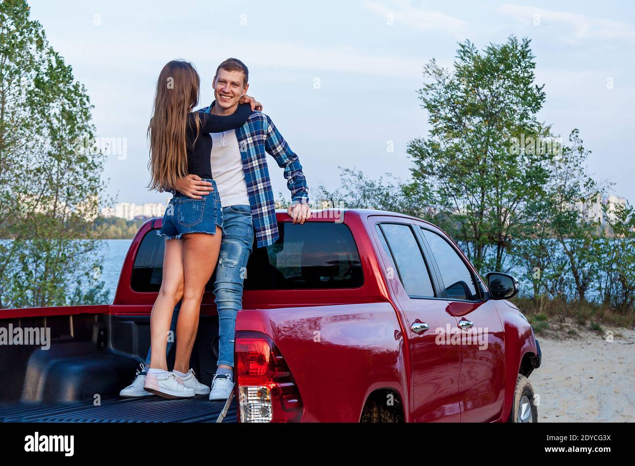 Happy young loving couple smiling and hugging in the back of a red car on the beach near the lake. Young couple hugs near the car. Stock Photo