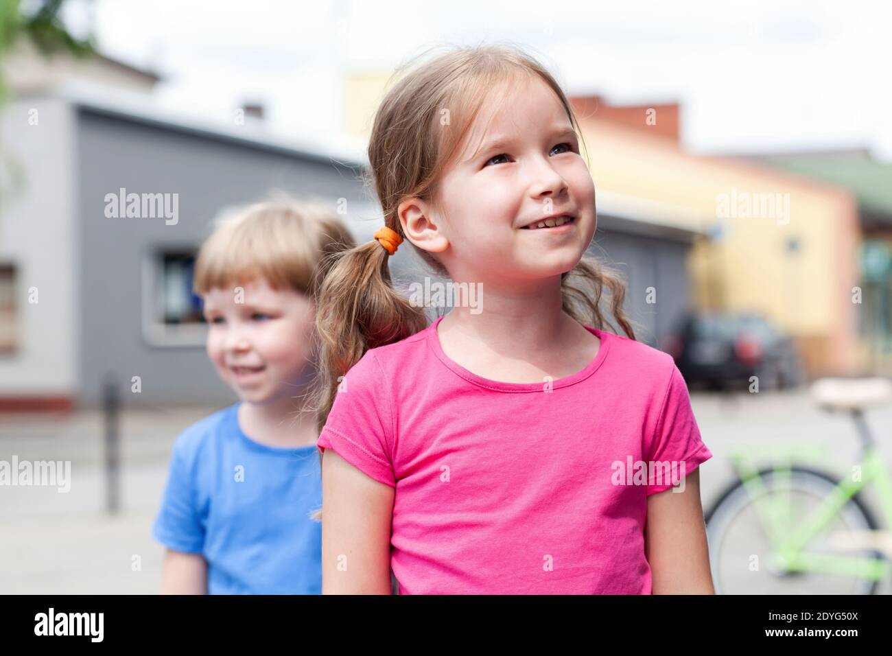 Happy, cheerful little school age girl with twin tails smiling, two sisters on the street together, one girl looking up at the parent, siblings Stock Photo