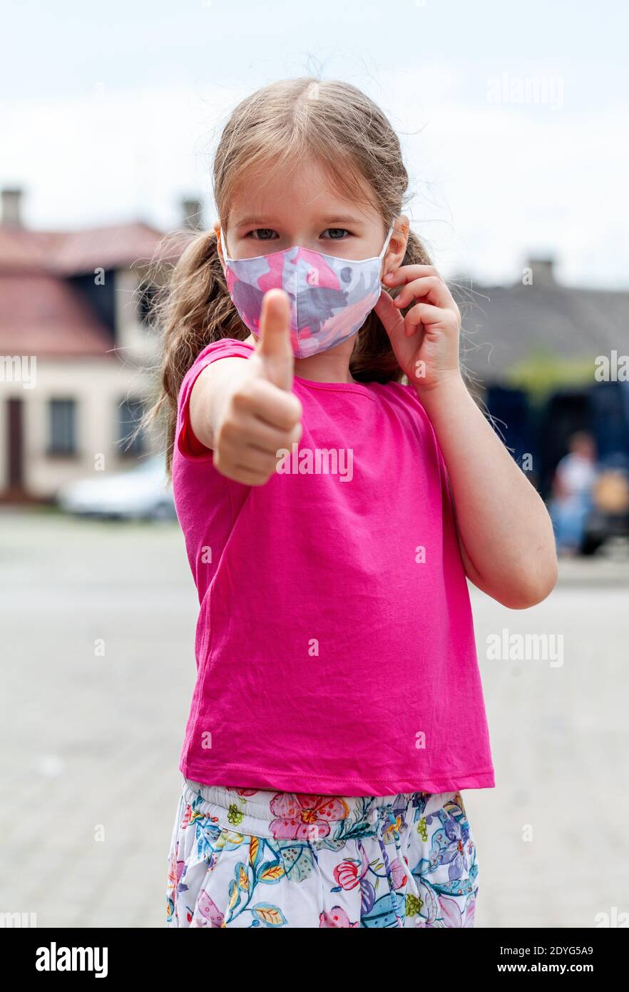 Little girl, young school age child wearing a colorful protective face mask showing thumbs up gesture on the street, portrait, closeup Stock Photo