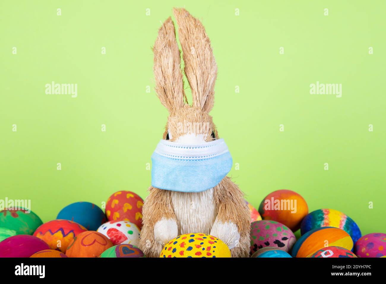 rabbit figurine with face mask between painted easter eggs before gren background, symbol for impact of corona virus on easter season Stock Photo