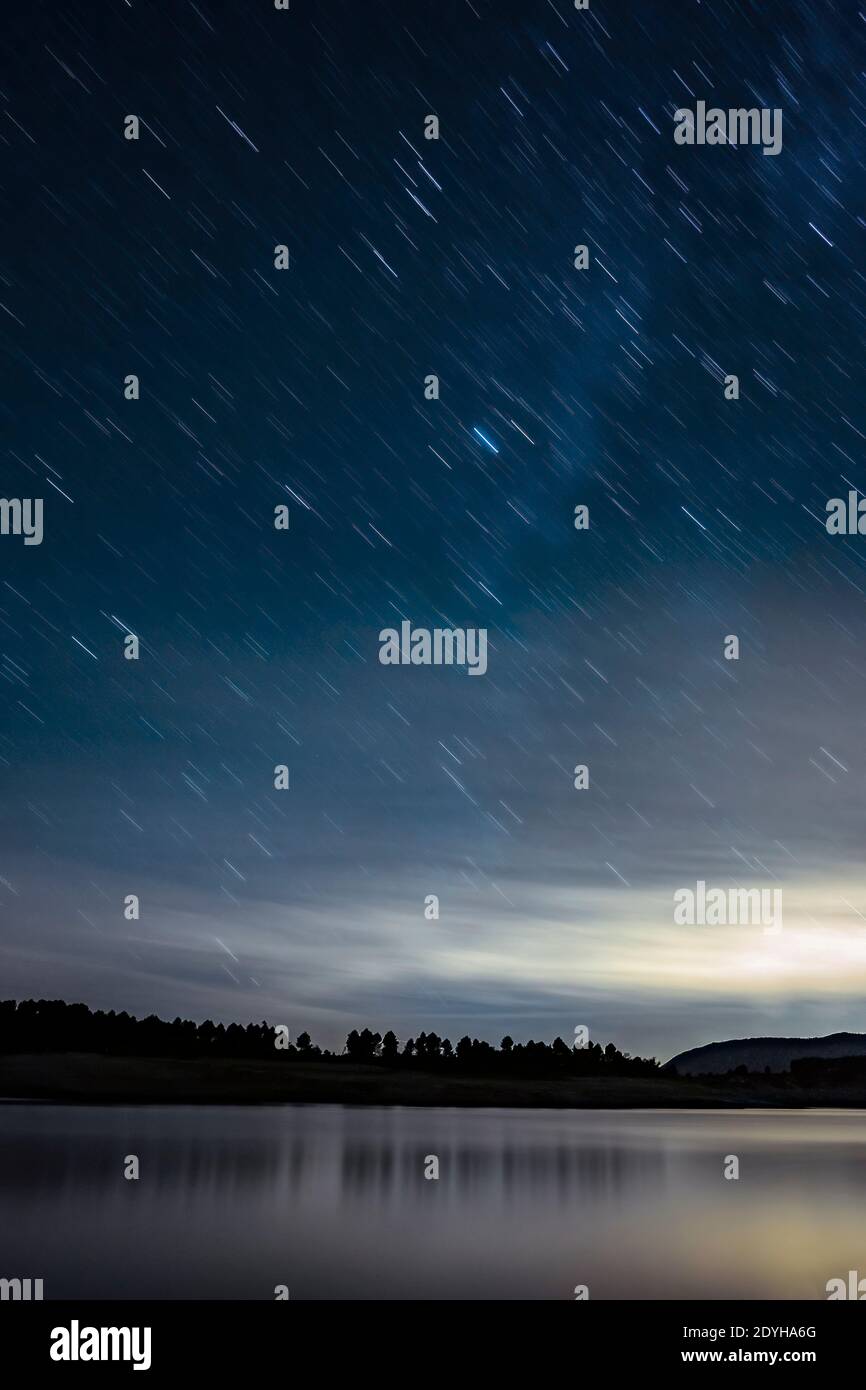 night landscape with a sky with stars trail, with the silhouette of a forest in the background and a lake in the foreground, vertical Stock Photo