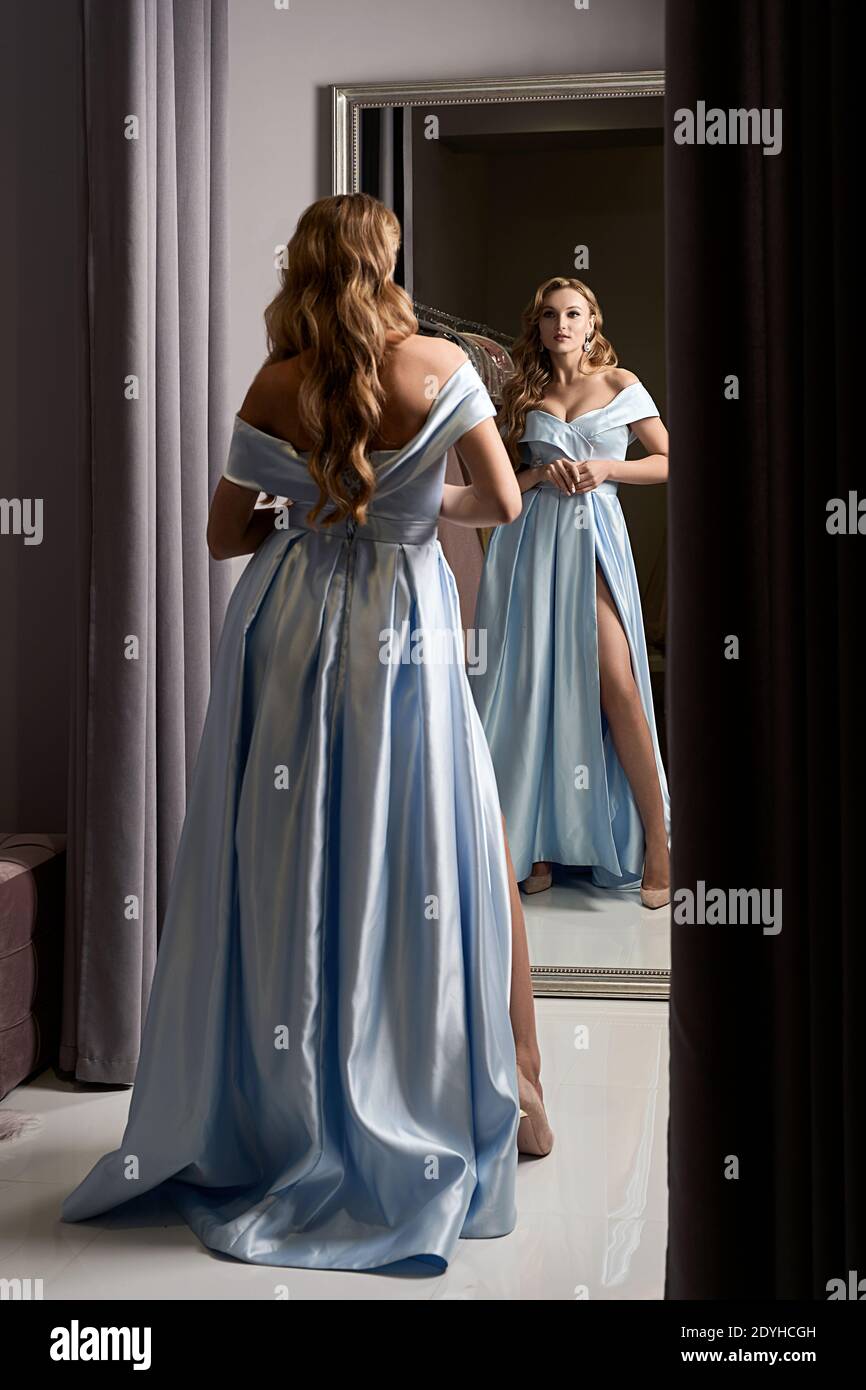 Young beautiful blonde girl wearing an off the shoulder full length sky blue satin slit prom ball gown. Model looking in mirror. Fitting room in dress Stock Photo Alamy