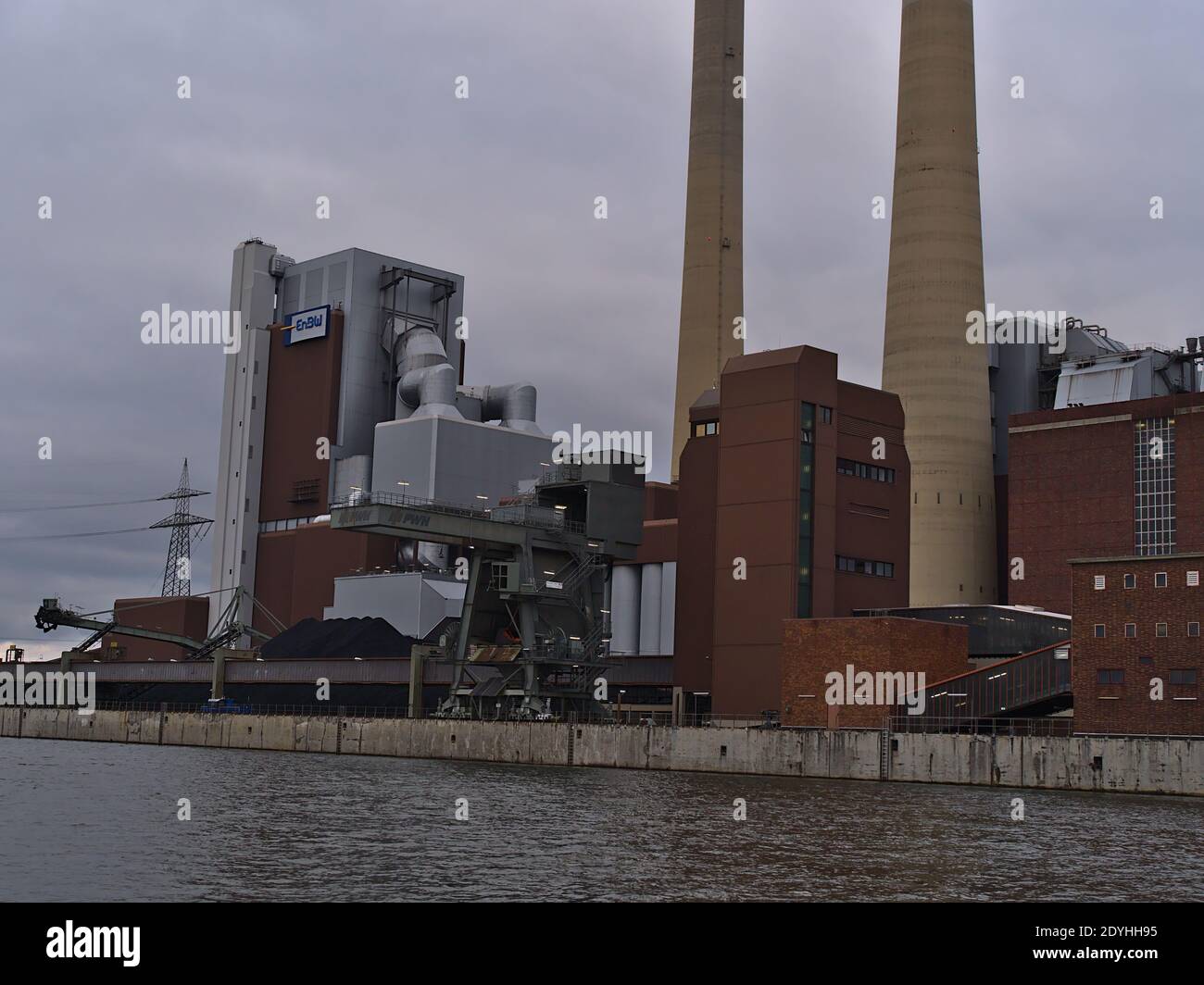 View of coal-fired Heilbronn Power Station operated by Energie Baden-Württemberg (ENBW) with heap of coal and port crane on th shore of Neckar river. Stock Photo