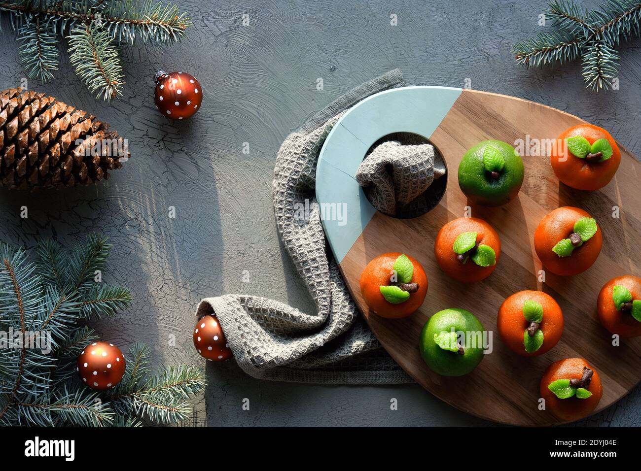 Marzipan sponge apples. Christmas dessert on wooden board. Cup of tea, fir twigs, red toys and pine cone. Top view on textile towel. Stock Photo