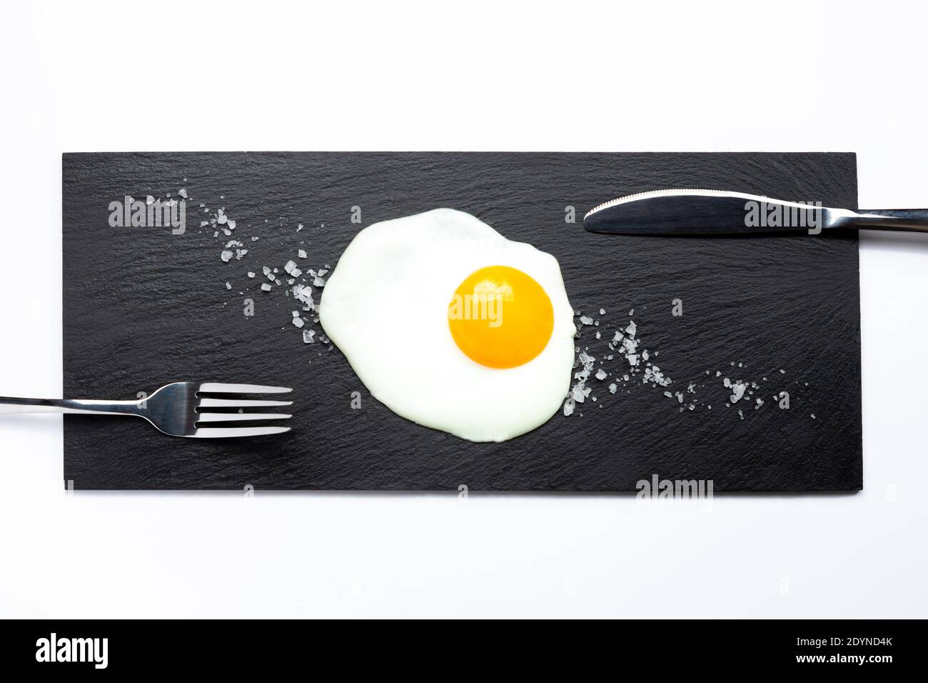 Fried egg on slate plate with knife, fork and salt on white background. Cooking and food concept. Stock Photo