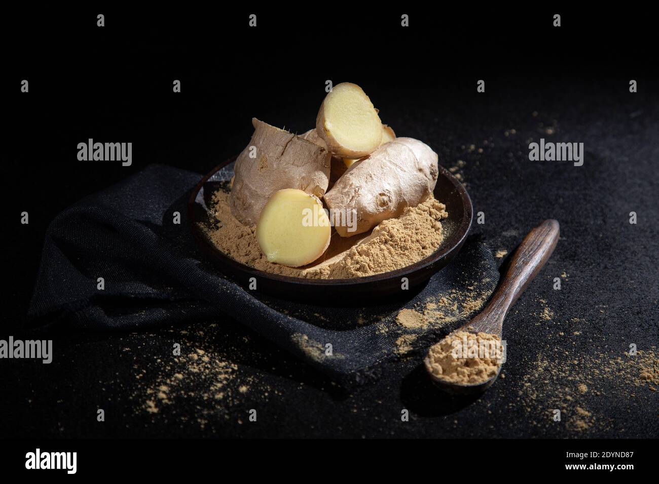 Pieces of ginger and ginger powder, in  wooden spoon and plate,  on black background. Stock Photo